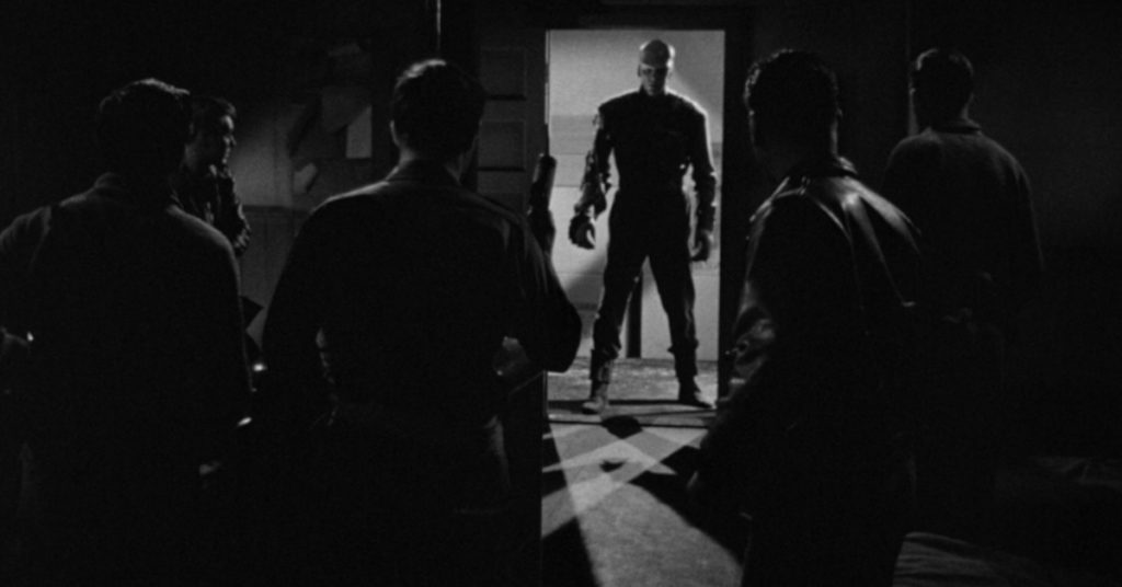 The Thing stands darkened in doorway as Captain Hendry and his men stand in shadows anticipating attack.