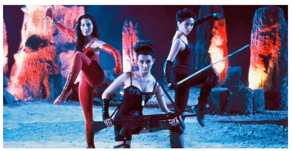 Michelle Yeoh, Maggie Cheung, and Anita Mui (pictured left to right) stare directly at the camera, ready for action in a nighttime scene from The Heroic Trio. They are all wearing sleeveless bodysuits and leggings with matching long gloves. Each holds a weapon in their hands.