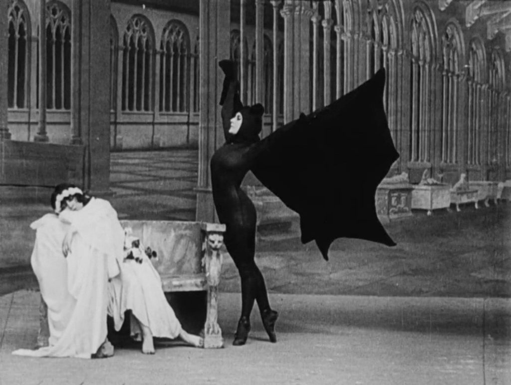 From Les Vampires (1915), the extravagant Irma Vep wears her signature black suit and wings while a woman sits passed out on a bench.