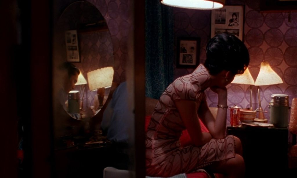 Maggie Cheung is looking away the camera, sitting on the edge of a bed and wearing an abstract printed pale pink cheongsam dress. Tony Leung is seen in the reflection of a mirror at a desk next to Cheung. 