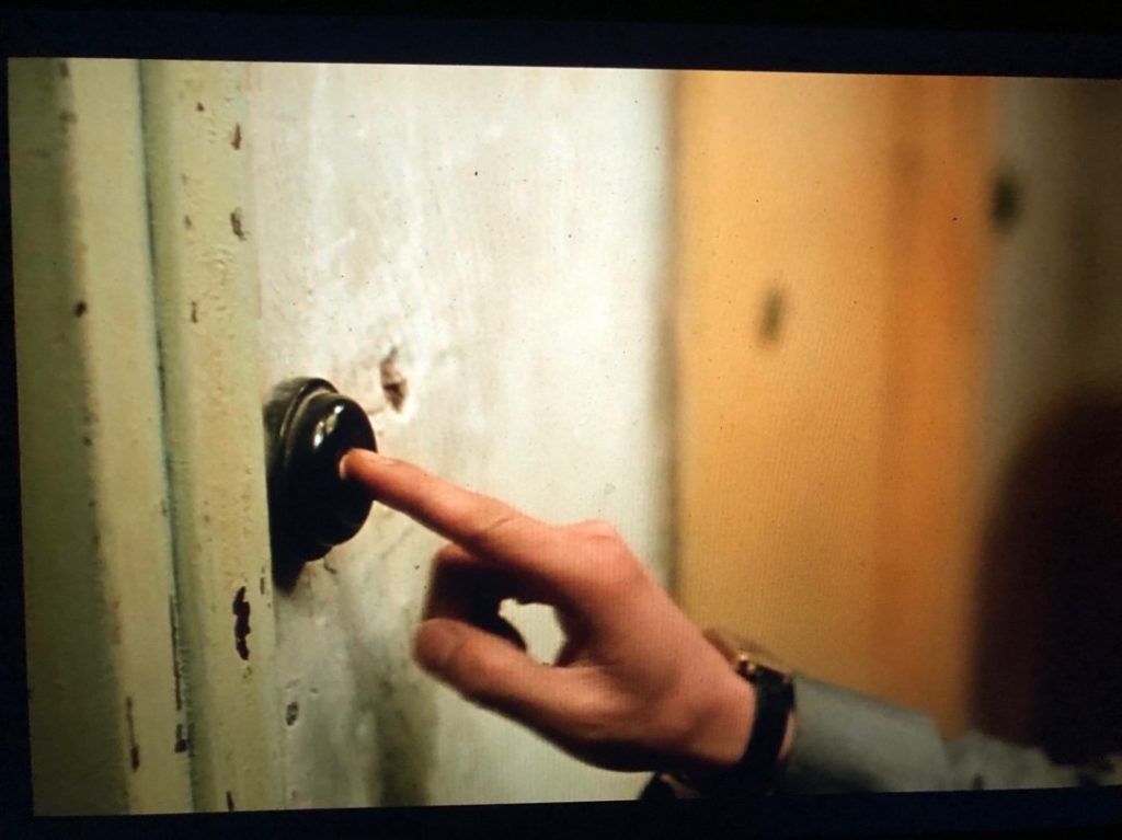 A man's right hand extends to press the button at the center of a black doorbell; the walls have paint peeling; he's wearing a grey suit and around his wrist is a gold watch with a black strap.