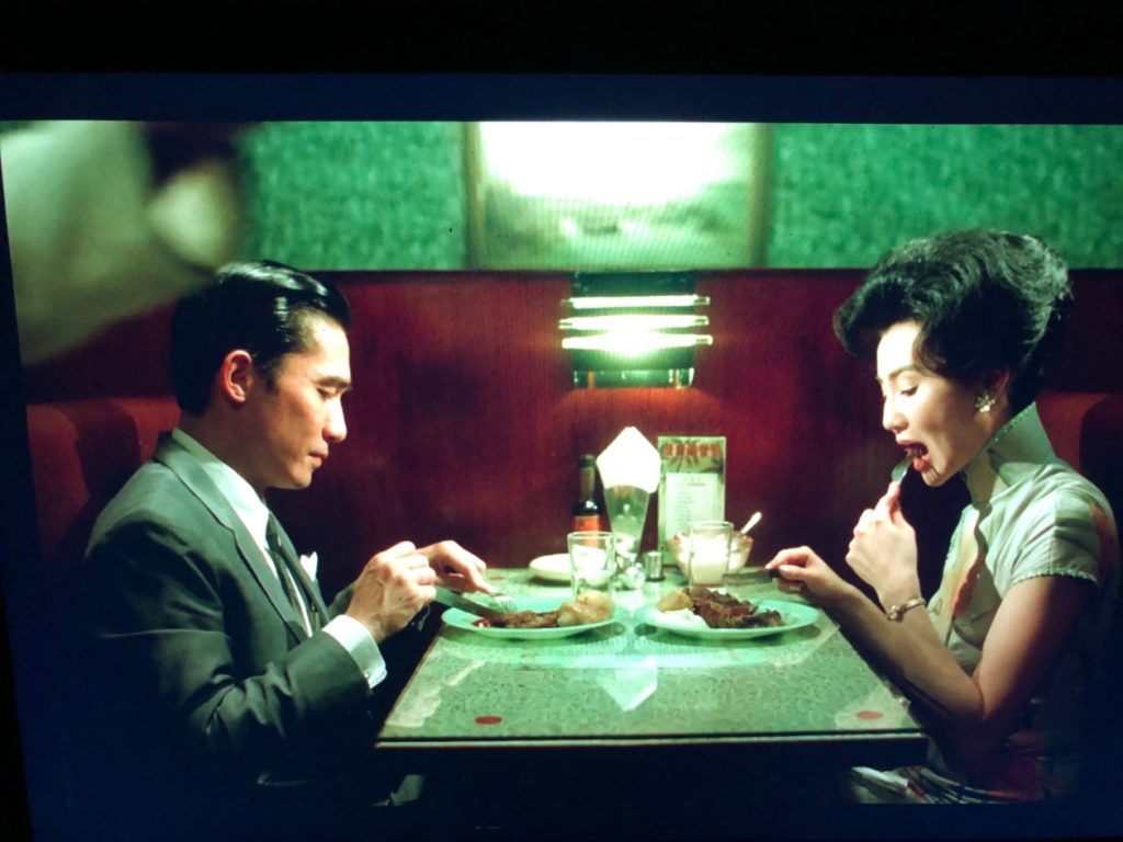 A man and a woman sit at a restaurant booth, each with a plate of steak and potatoes, eating dinner together. He wears a grey suit and tie, she wears a patterned dress.