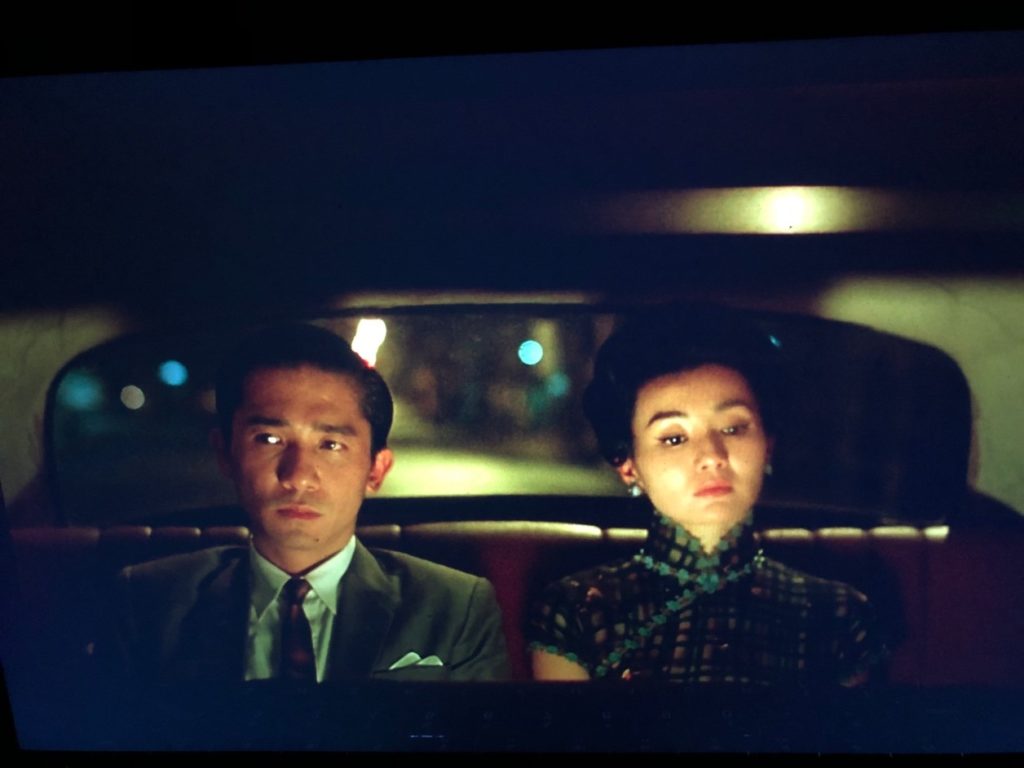 A man and a woman sit in the back seat of a taxi, facing forward, not looking at each other. Through the back window we can see the night street, illuminated by city lights.