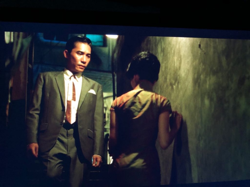 A man in a grey suit and rust & white tie walks down a narrow staircase while a woman in a floral print dress walks up the same stairwell; grey concrete walls, a street lamp overhead.