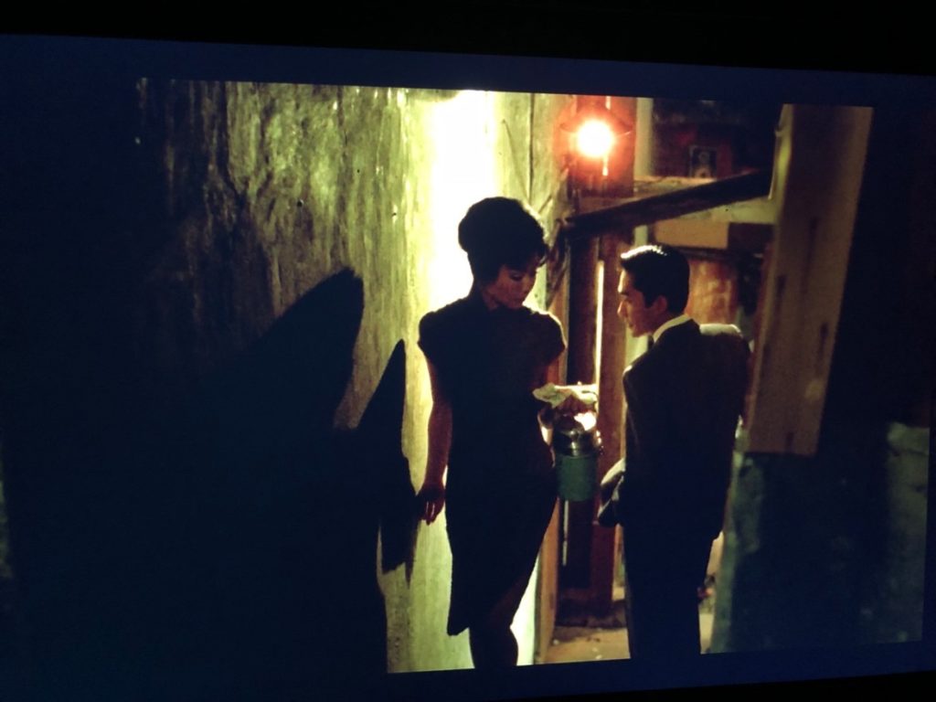 A man in a grey suit and rust & white tie walks down a narrow staircase while a woman in a floral print dress walks up the same stairwell; grey concrete walls, a street lamp overhead.