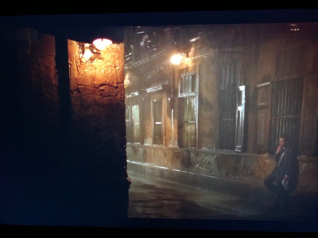 ABOVE: A man in a grey suit smokes a cigarette while leaning against the window of a grey building in the rain, a street lamp in the foreground, another closer to him. BELOW: A woman in a floral dress in the same exact position in the scene, a few moments later. She leans against a grey building in the rain, a street lamp in the foreground, another closer to her.