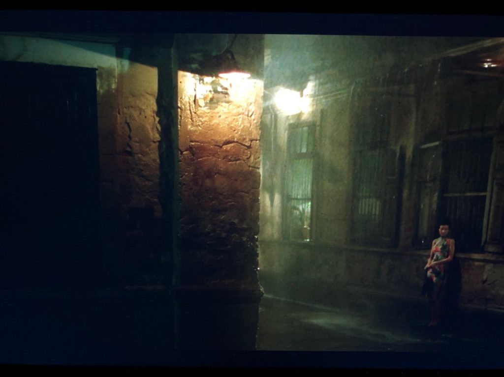 ABOVE: A man in a grey suit smokes a cigarette while leaning against the window of a grey building in the rain, a street lamp in the foreground, another closer to him. BELOW: A woman in a floral dress in the same exact position in the scene, a few moments later. She leans against a grey building in the rain, a street lamp in the foreground, another closer to her.