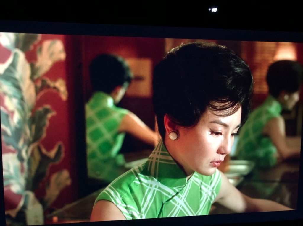 A woman sits at her dressing table in a green dress and white oval earrings, downcast after crying. You can see her from two additional angles in the mirror behind her.