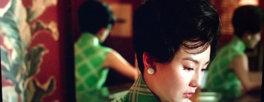 A woman sits at her dressing table in a green dress and white oval earrings, downcast after crying. You can see her from two additional angles in the mirror behind her.