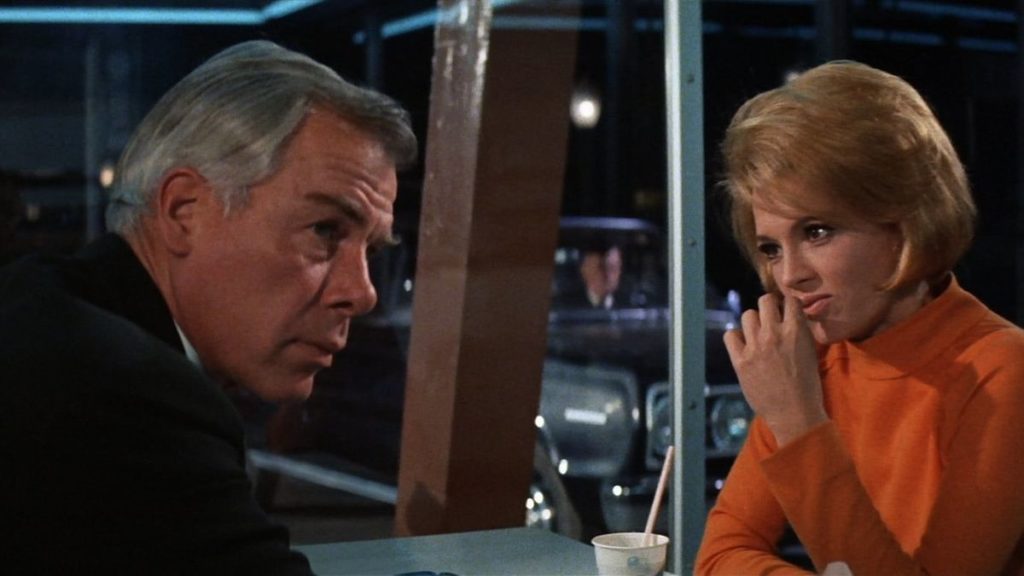 A man (Lee Marvin) and a woman in an orange shirt (Angie Dickinson) sit at a diner table at night, a man in a car waiting in the parking lot visible through the windows behind them. 