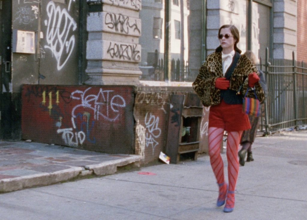 A woman (Parker Posey) wearing blue high heels, red stockings, a red skirt, red gloves, leopard-print jacket, and sunglasses walks down a sidewalk with buildings littered with graffiti behind her. 