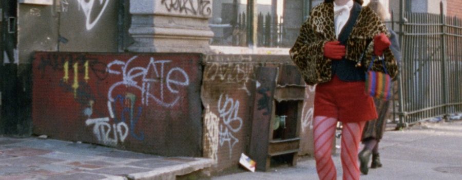 A woman (Parker Posey) wearing blue high heels, red stockings, a red skirt, red gloves, leopard-print jacket, and sunglasses walks down a sidewalk with buildings littered with graffiti behind her.