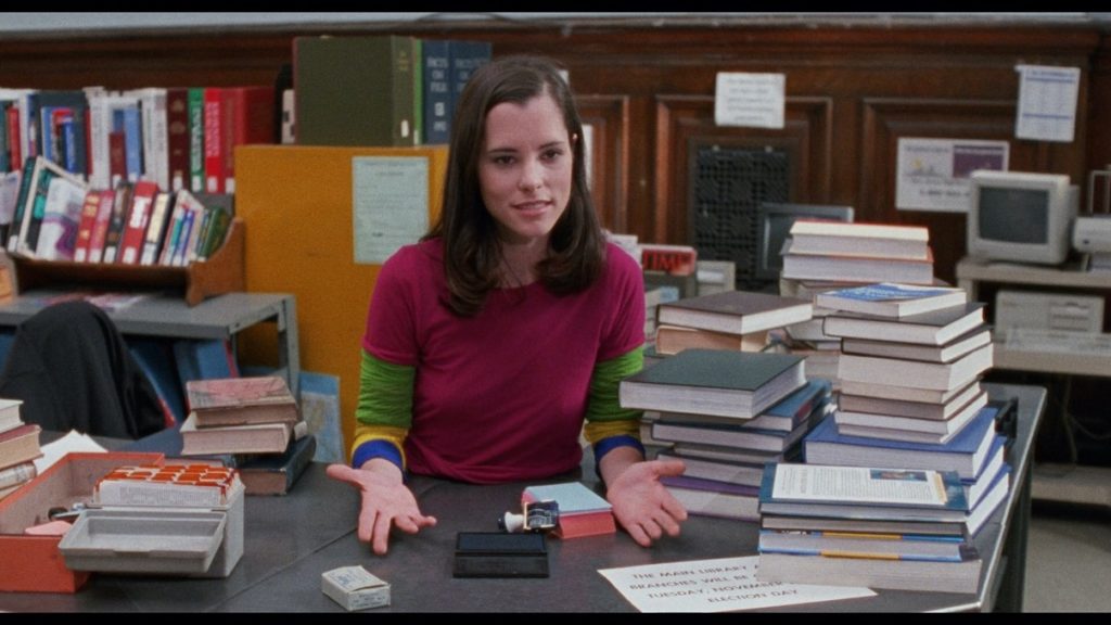 9. A photo of Parker Posey in a library wearing a bright pink crew neck t-shirt over a green t-shirt and she is surrounded by stacks of books.
