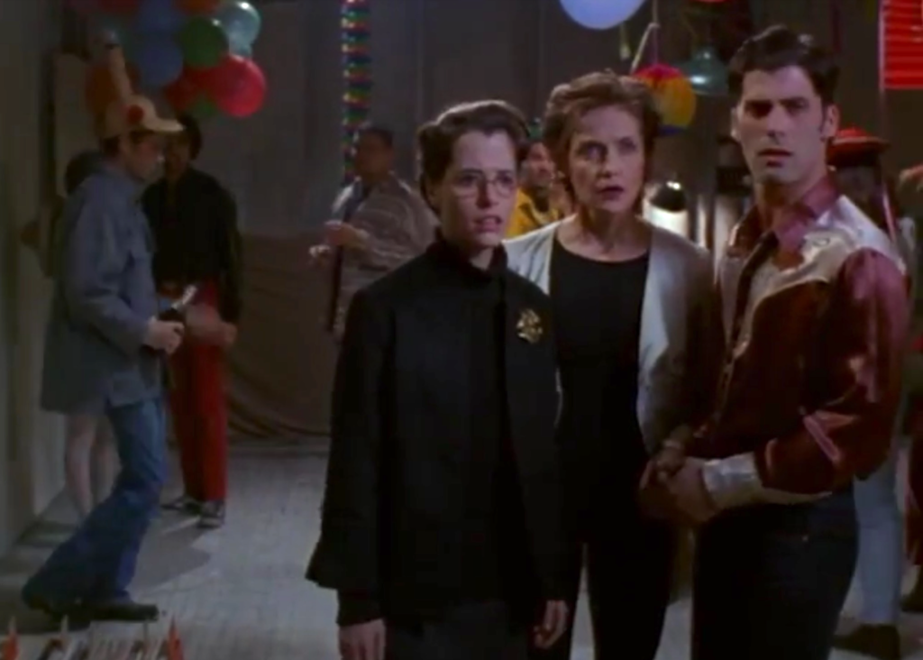 11. A photo of Parker Posey, Donna Mitchell, and Anthony DeSando at a party. Posey is wearing glasses and a black suit with a broach. Mitchell is wearing a white jacket over a black top and pants. DeSando is wearing a gold and burgundy western shirt.
