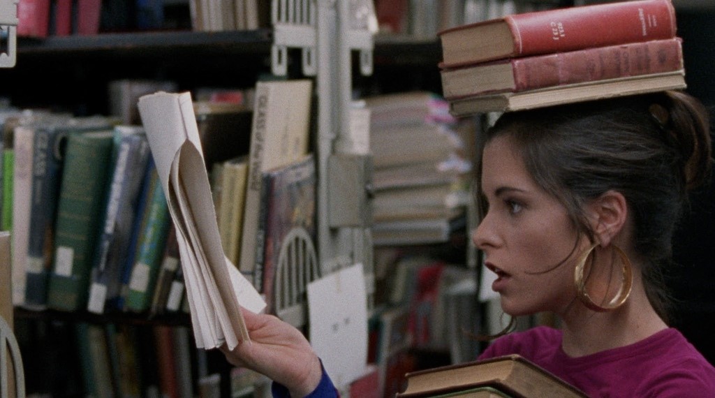 3. Parker Posey wearing a bright pink, crew neck t-shirt with large good hoop earrings in a library holding books in one hand and a white sheet of paper in the other. She also has books stacked on her head.
