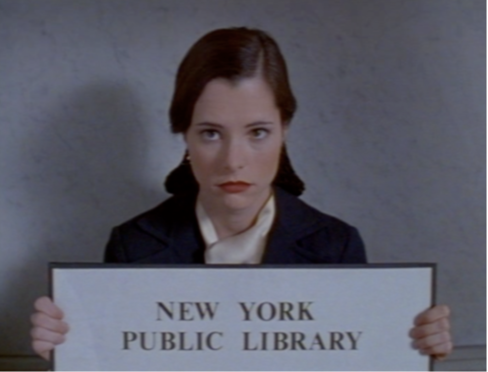 4. Parker Posey in a white blouse and black jacket holding a black and white sign that says "New York Public Library".
