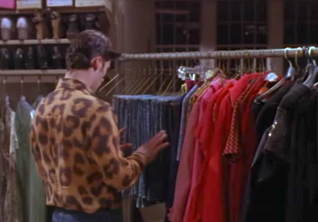 8. A photo of a man wearing a leopard print shirt while looking at a rack of clothing that includes several pairs of jeans.
