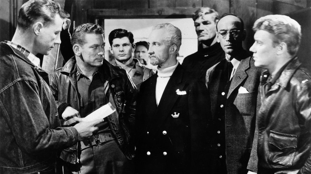 A blond man wearing a dark coat stands in the middle of a group of men who look at him suspiciously. 