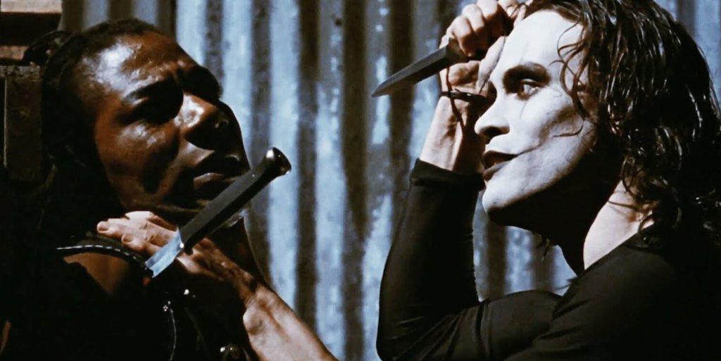 On the right, Brandon Lee as Eric Draven threatens a gang member (posed on the left) with a knife; there is already a knife imbedded in the gangsters's shoulder.