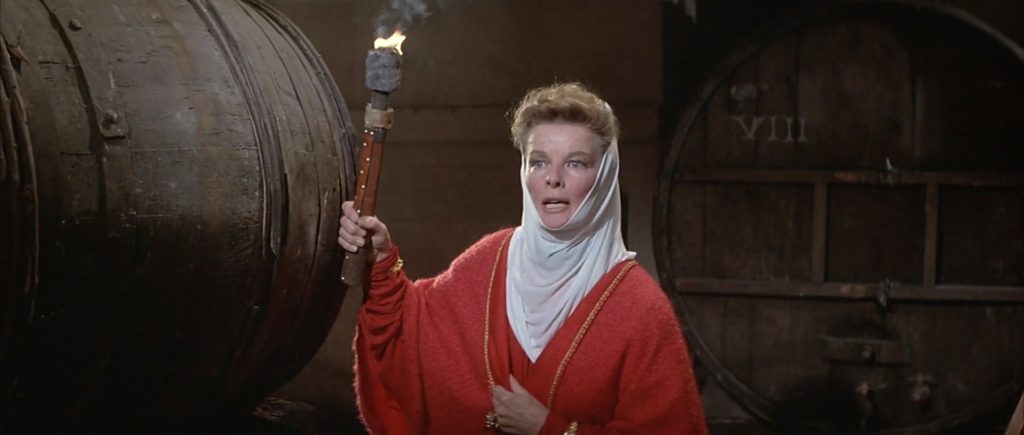 Queen Eleanor in a red dress holds a candle beside large barrel in the cellar.