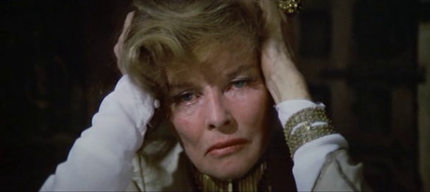 Eleanor (Katharine Hepburn) clutches her head in distress. She is wearing a white dress shirt with golden bracelets adorning each arm.