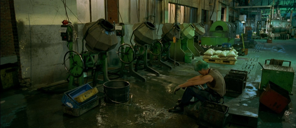 In a dimly lit factory, a green haired man sits, defeated.