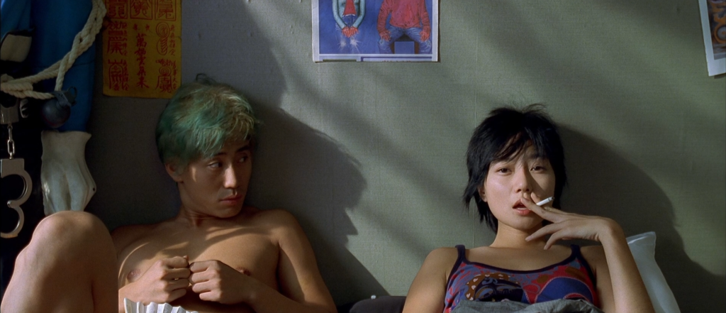 A green haired man and his dark-haired girlfriend talk in bed. she is smoking a cigarette, he is looking at her.