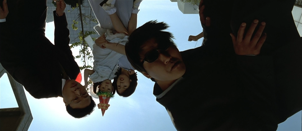 Four members of a family bend their heads to look upside down at something out of frame. The man closest to the camera is twisted around and is dressed totally in black. Two of the figures in the back are dressed in white, one is a child and one is an adult woman. The last family member, directly on the left, is dressed in black.