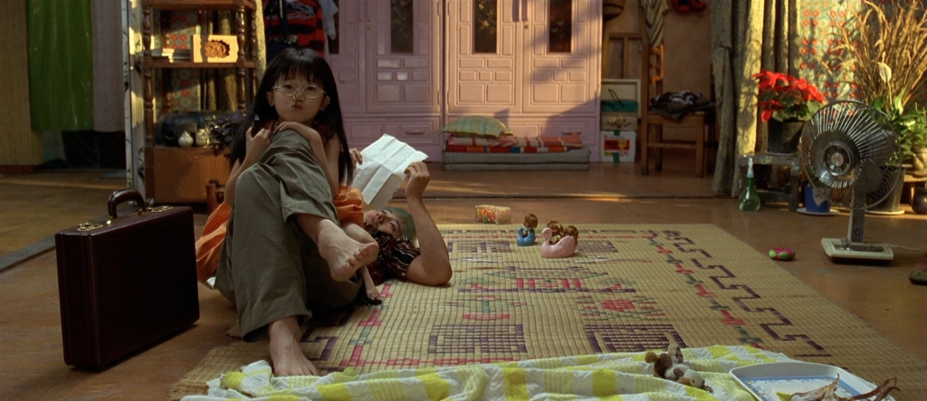 In a messy room, a girl rests on a man's lap while he reads a letter.