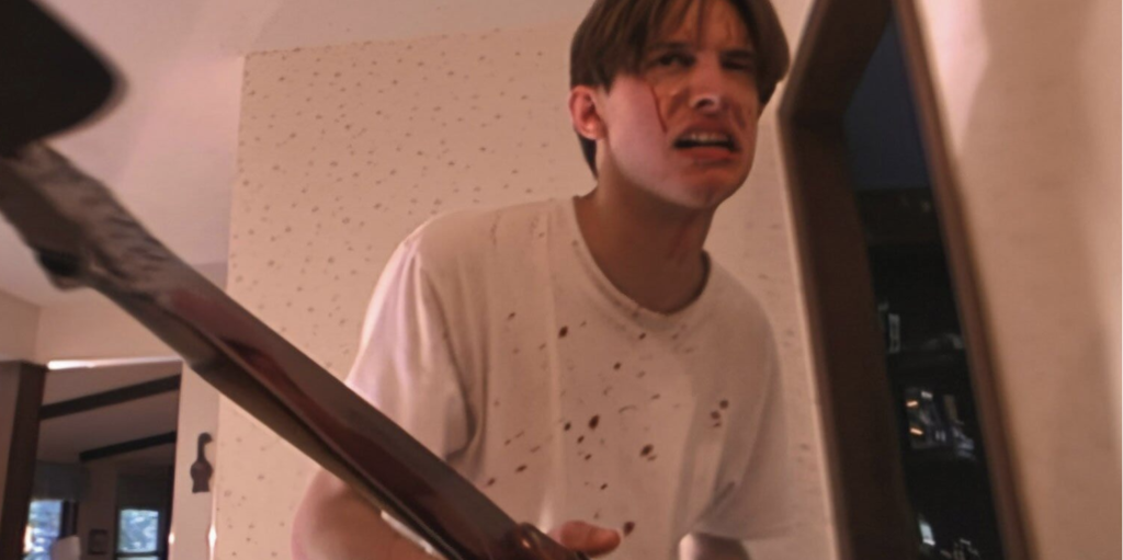 A man stands in a white hallway, wearing a white t-shirt that is splattered with blood. Blood streaks down his cheeks. He is holding an ax, which is dangerously close to the camera.