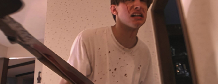A man stands in a white hallway, wearing a white t-shirt that is splattered with blood. Blood streaks down his cheeks. He is holding an ax, which is dangerously close to the camera.