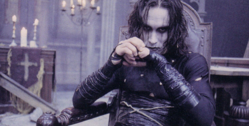 Brandon Lee is dressed in black and has white face makeup. He is sitting in a dilapidated, gothic room.
