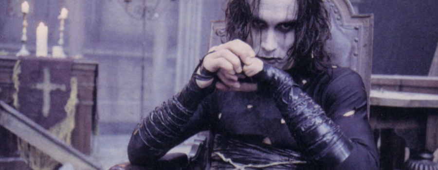 Brandon Lee is dressed in black and has white face makeup. He is sitting in a dilapidated, gothic room.