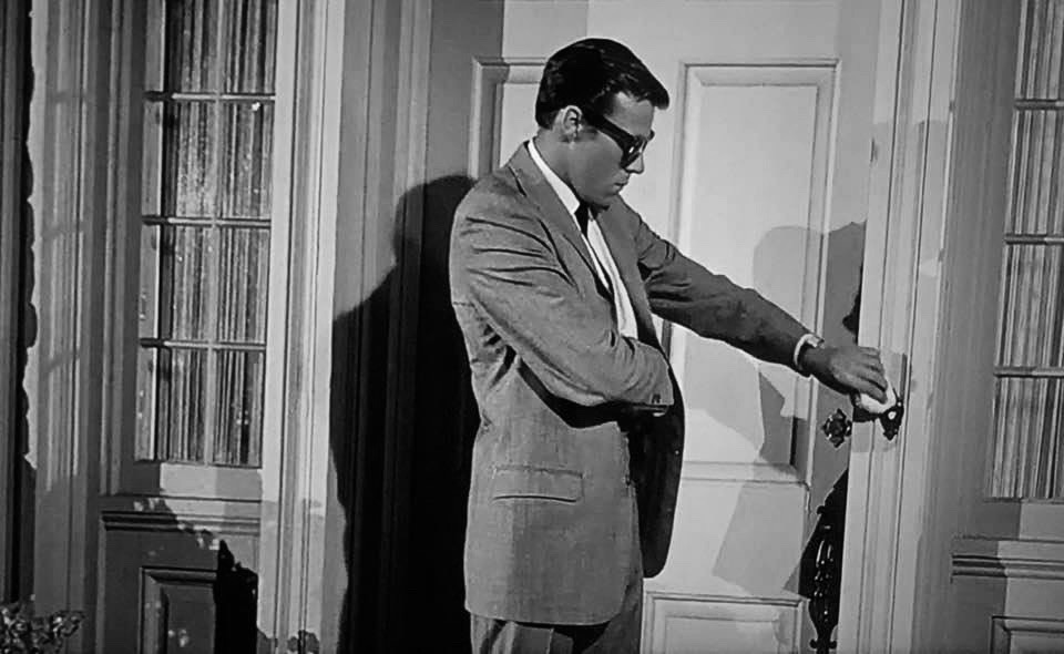 A man wearing a suit and a pair of sunglasses puts his right hand in his pocket and has his left hand on a door.