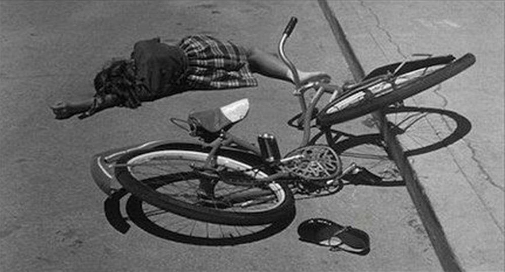 A girl, dead, is lying on the pavement. Her broken bicycle is in front of her.