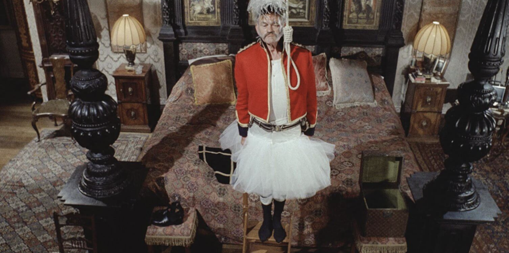 In the middle of a bedroom, a man dressed in a tutu and a red jacket is standing on a stool. A noose hangs in front of him.