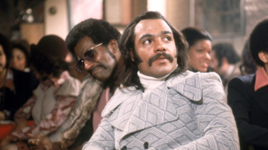 Actor Carl Lee, a Black man wearing sunglasses and a fur coat, and actor Ron O'Neal, a Black man with long hair, a mustache, a white turtleneck and a gray and white peacoat are seen in still from the film Super Fly that depicts a nightclub scene with numerous extras in the background.