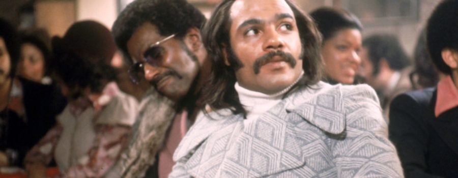 Actor Carl Lee, a Black man wearing sunglasses and a fur coat, and actor Ron O'Neal, a Black man with long hair, a mustache, a white turtleneck and a gray and white peacoat are seen in still from the film Super Fly that depicts a nightclub scene with numerous extras in the background.