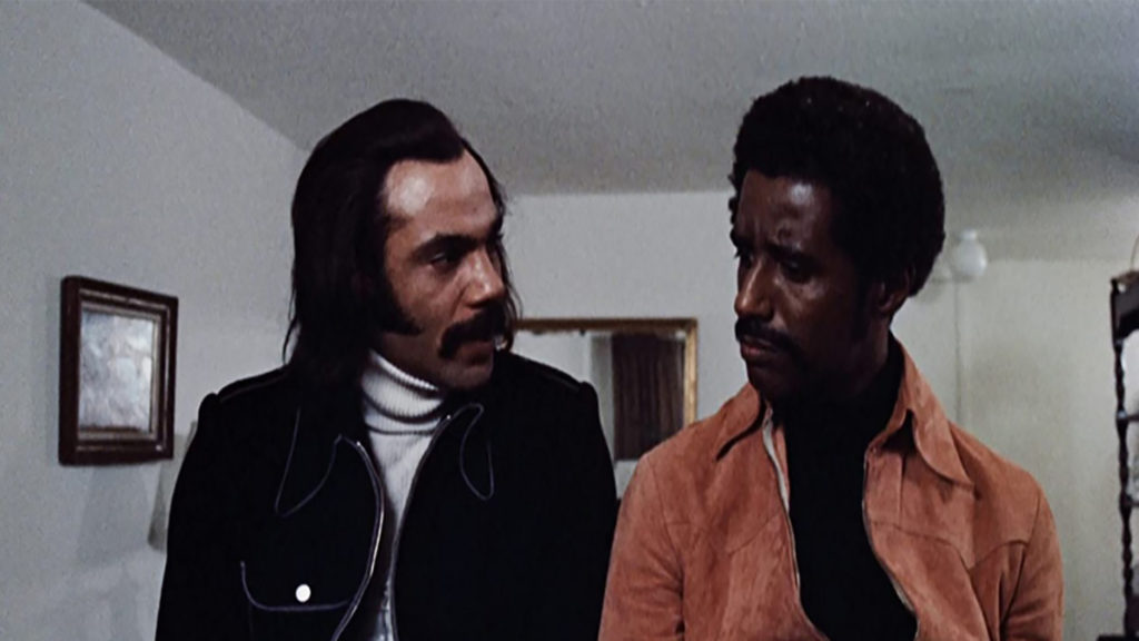 A still from the film Super Fly showing Ron O'Neal, a Black man wearing a white turtleneck and a black jacket speaking to Carl Lee, a Black man wearing a black shirt and a suede jacket.