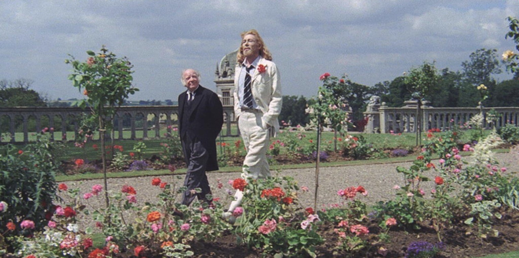 Two men walk through a botanical garden. The one on the left is dressed in black, while the one on the right is wearing a white suit.