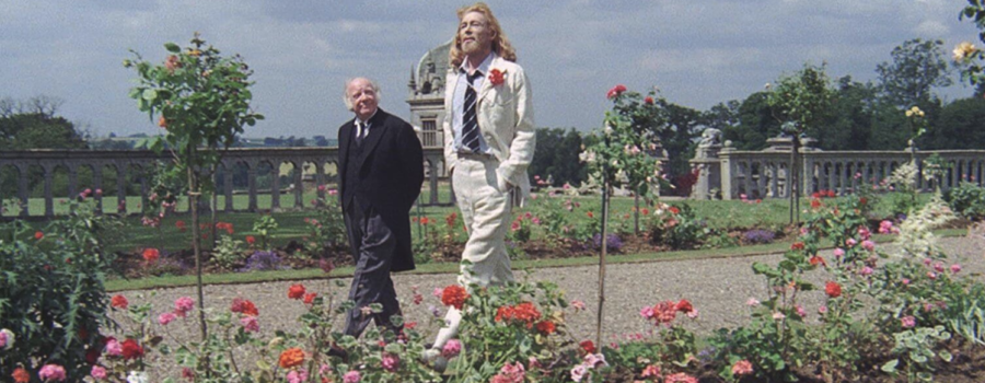 Two men walk through a botanical garden. The one on the left is dressed in black, while the one on the right is wearing a white suit.