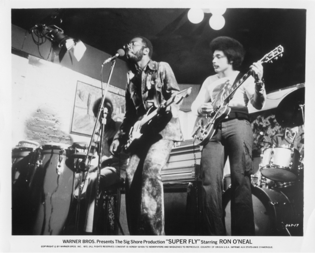 A band of men are playing a song in a well-lit room. The photo is in black and white. At the bottom of the image is text that reads "Warner Bros presents the Sig Shore production "SUPER FLY" starring RON O'NEAL