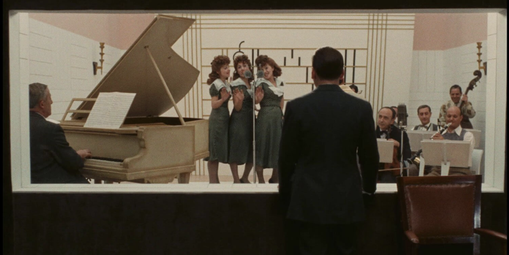 A man stares at a group of women singing with a band. On the left, an older man plays an ivory colored piano. Next to him are three red headed women wearing green dresses, singing. To their right is a small orchestra.