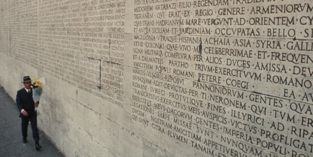 In the bottom left corner, a man in a suit carries a bouquet of yellow flowers. He is walking past a large brick wall with words in Latin carved on it.