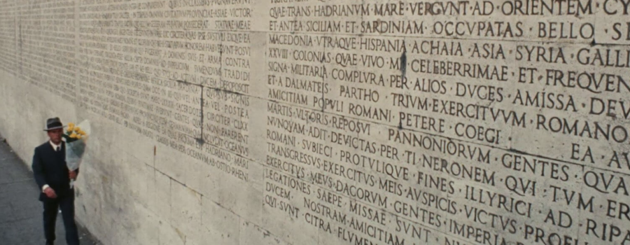 In the bottom left corner, a man in a suit carries a bouquet of yellow flowers. He is walking past a large brick wall with words in Latin carved on it.