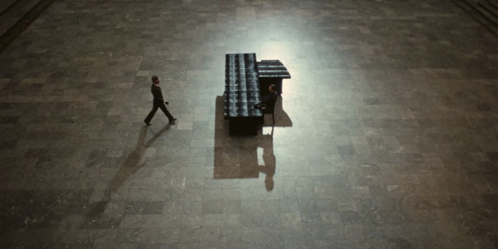 In an empty room, a man sits at a large desk. Another man in a hat approaches him from the left.