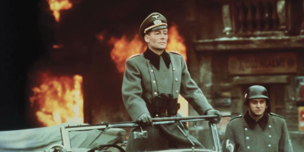 A man in an olive green Nazi uniform stands upright in a military vehicle, wearing a pair of binoculars around his neck. To his right, there is a man in a small bucket hat. Behind them, there are several fires raging.