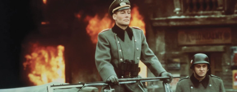 A man in an olive green Nazi uniform stands upright in a military vehicle, wearing a pair of binoculars around his neck. To his right, there is a man in a small bucket hat. Behind them, there are several fires raging.