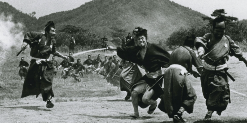 In a black and white image, an army of men attack two samurai. There are wooded mountains in the background of the image.