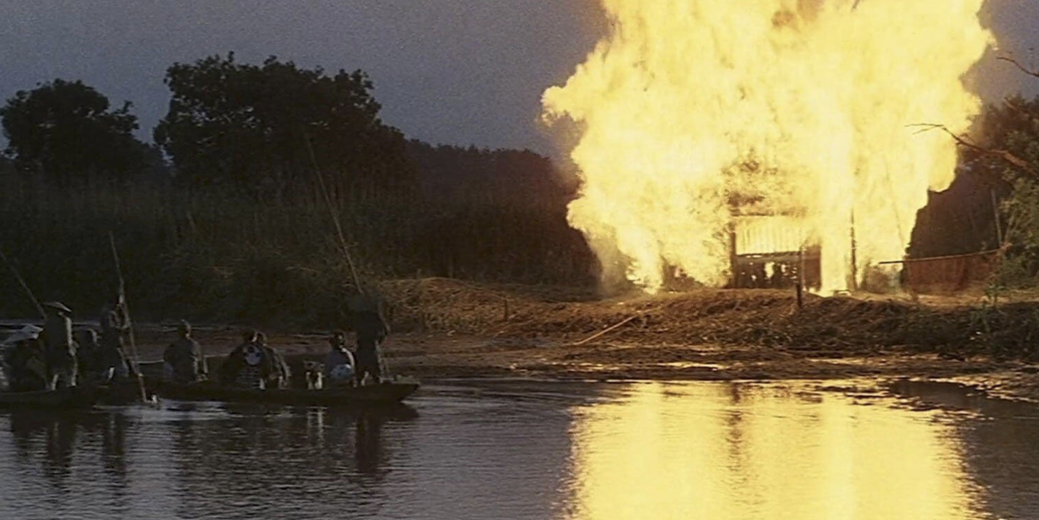 On the left, a group of people approach a riverbank on two boats. On the right, a building is blazing in a roaring fire.